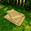 Folded Madurkathi grass mat with ethnic border placed on green grass in a natural outdoor garden setting.
