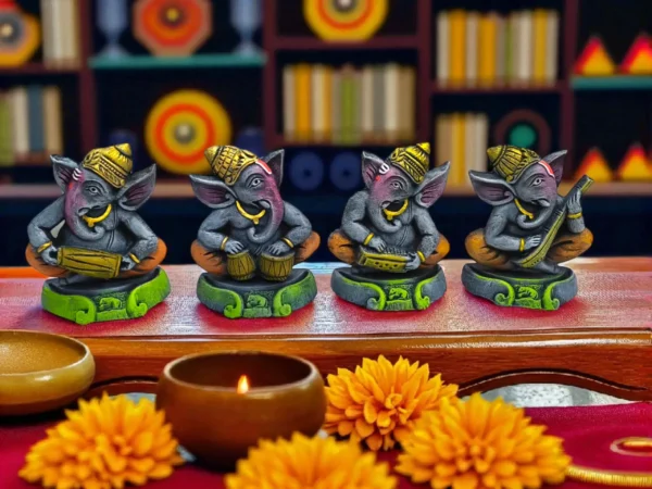 Handmade grey burnt clay Ganesha musician figurines set of four with yellow-orange details and green bases, displayed on a wooden surface with marigold flowers and a lit diya, against a festive background.