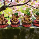 Handmade multicolor burnt clay Ganesha musician figurines set of four with green caps and vibrant detailing, placed on a moss-covered stone surface with blooming pink flowers in the background.