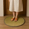 A person standing on a round, green natural grass mat placed on a wooden floor in front of a door, wearing brown slippers and a light floral dress.