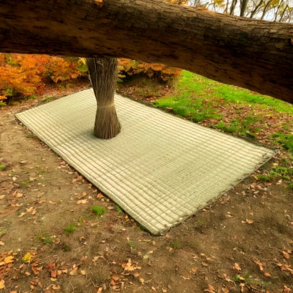 Handwoven Madurkathi grass mat laid on the ground outdoors beneath a tree with a tied bundle of rivergrass in the center.