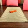 Handwoven Madurkathi grass mat placed indoors on wooden flooring with red cushions and an orange cloth.