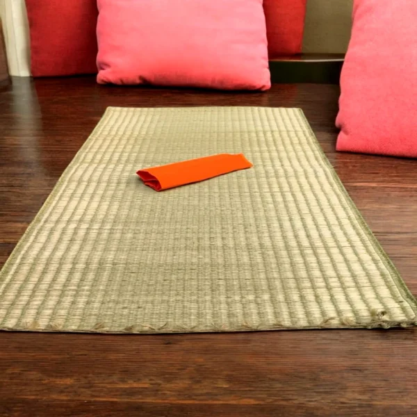 Handwoven Madurkathi grass mat placed indoors on wooden flooring with red cushions and an orange cloth.