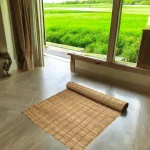 Handmade river grass mat partially rolled on a clean indoor floor with a scenic view of green fields through large windows – eco-friendly and foldable mat, size 3x7 ft.