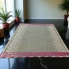 Handmade traditional mat with intricate geometric patterns and pink border, placed in a modern indoor setting with black marble flooring and potted plants.