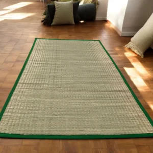 Natural handwoven river grass mat with green border placed in a cozy indoor room with wooden flooring.