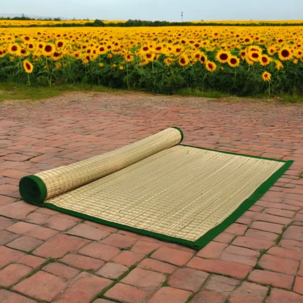 Handwoven natural river grass mat with green border partially rolled out on brick pavement near a blooming sunflower field.