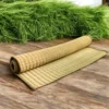 Rolled handmade Madurkathi grass mat placed on a wooden floor with fresh green rivergrass in the background.