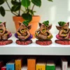 Four colorful handcrafted clay Ganesha statues, each playing a musical instrument, placed on a white shelf with books and a potted plant in the background.