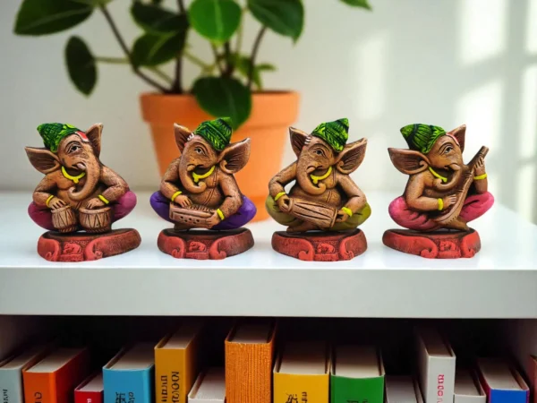 Four colorful handcrafted clay Ganesha statues, each playing a musical instrument, placed on a white shelf with books and a potted plant in the background.