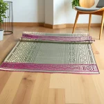 Partially rolled handmade grass mat with traditional patterns and pink border, displayed on a light wooden floor beside a modern chair and indoor plants.