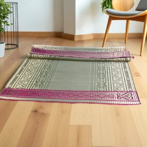 Partially rolled handmade grass mat with traditional patterns and pink border, displayed on a light wooden floor beside a modern chair and indoor plants.