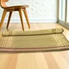 Partially rolled Madurkathi grass mat with ethnic border placed on a light wooden floor near a wooden chair and glass door.