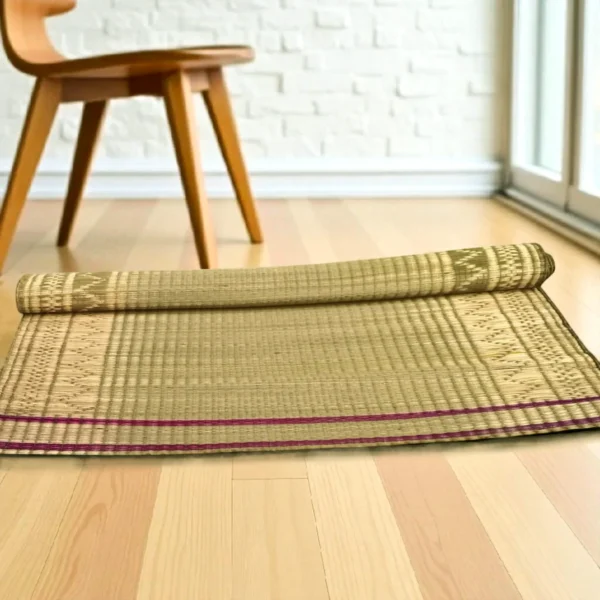 Partially rolled Madurkathi grass mat with ethnic border placed on a light wooden floor near a wooden chair and glass door.