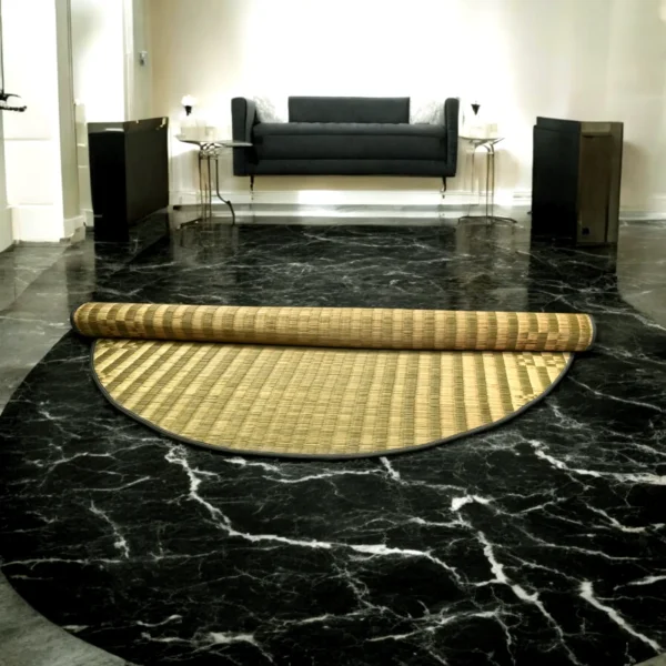 Partially unrolled 4ft round Madurkathi river grass mat on a black marble floor in a modern, elegant room with a black sofa