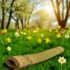 Rolled Madurkathi grass mat on green grass in a sunny park with blooming yellow, white, and pink flowers.