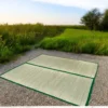Two large handwoven river grass mats with green borders laid out on a gravel path surrounded by lush green fields.