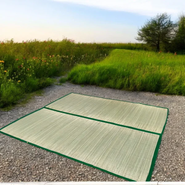 Two large handwoven river grass mats with green borders laid out on a gravel path surrounded by lush green fields.