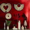 A stylish wall decorated with three natural handwoven grass wall hangings—heart, round, and fan shapes—set against a rich red background with modern vases and plants below.
