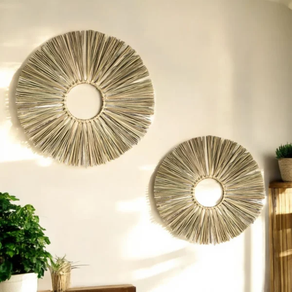 Two round sunburst-style wall hangings made from natural grass straw, mounted on a cream-colored wall with sunlight casting warm shadows and plants nearby.