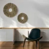 Two handcrafted round sunburst wall hangings made from natural grass, displayed on a white wall above a wooden floor and modern lounge chair.