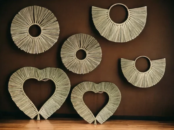 A set of six handcrafted wall hangings in heart, round, and fan shapes made from natural straw, displayed against a deep brown wall with wooden flooring.