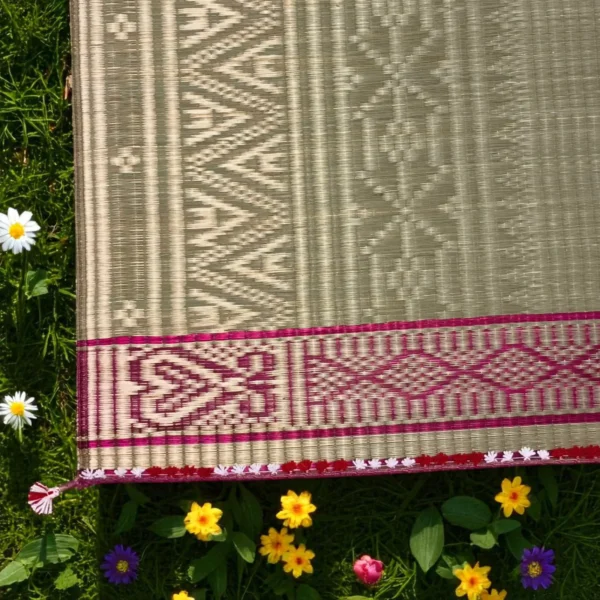 Close-up of a traditional handwoven river grass mat with intricate tribal patterns and a pink-maroon border, placed on green grass with colorful flowers around.