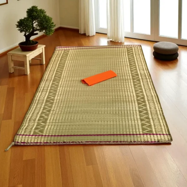 Madurkathi grass mat with ethnic border laid on a wooden floor in a serene indoor room with bonsai and cushions.