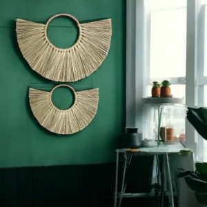 Two handmade half-moon wall hangings made of natural grass, displayed on a green wall in a boho-style room with indoor plants and natural light.