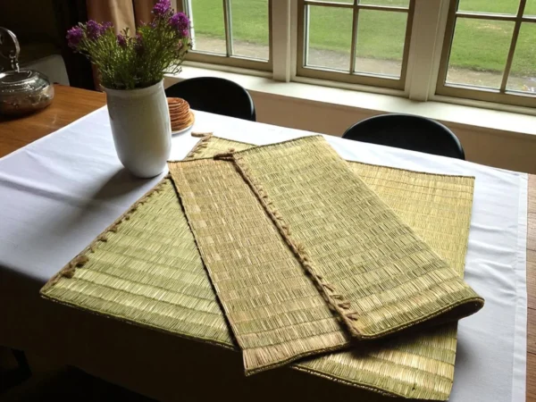 Folded natural grass mats placed on a dining table with a flower vase near a window.