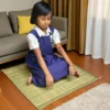 A schoolgirl sitting on a natural grass mat in a living room setting.