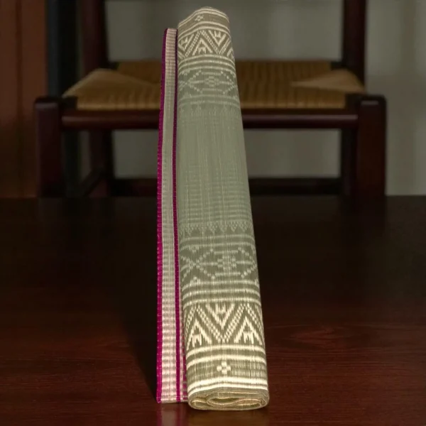 Rolled-up traditional handwoven grass mat with pink border and intricate ethnic patterns, standing upright on a wooden surface.