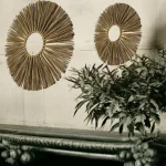 Two handcrafted round wall hangings made from natural straw, displayed on a rustic white wall above a traditional wooden bench and leafy plant decor.
