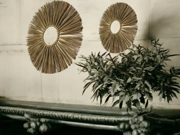 Two handcrafted round wall hangings made from natural straw, displayed on a rustic white wall above a traditional wooden bench and leafy plant decor.