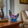 Handmade burnt clay Krishna face decorative statue with peacock feather, placed on a wooden side table beside books and a flower vase in a cozy living room.