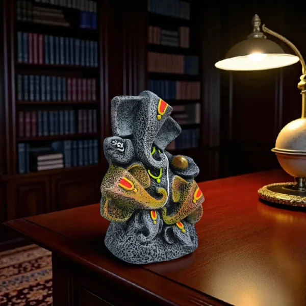 Handmade burnt clay Lord Ganesha statue in black terracotta finish with colorful accents, placed on a wooden desk with a lamp and bookshelf background.
