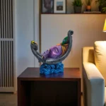 Handcrafted colorful clay Krishna idol playing flute on crescent moon, placed on wooden table in living room décor setup.