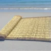 Handwoven natural Kusha grass mat placed on a sandy beach near the sea — eco-friendly mat ideal for yoga, meditation, or puja.