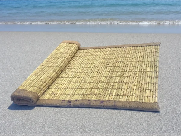 Handwoven natural Kusha grass mat placed on a sandy beach near the sea — eco-friendly mat ideal for yoga, meditation, or puja.
