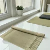 Partially rolled handmade reed grass (Madurkathi) mat placed on a white table near a window with a plant and other mats.