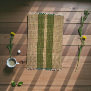 Handwoven natural grass table mat with green and beige stripes placed on a wooden surface with sunlight shadows, surrounded by flowers and a cup.