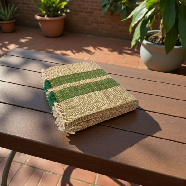Folded handwoven green and natural-striped grass mat placed on an outdoor wooden bench in a sunny patio with plants in the background.