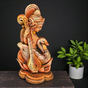 Handcrafted terracotta Goddess Saraswati statue holding a veena, seated with a swan, placed on a wooden surface against a dark background.
