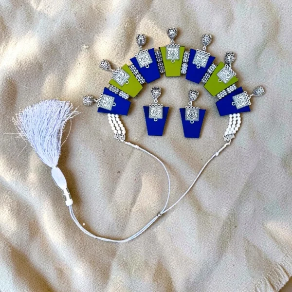 Handmade fabric necklace set with blue and green panels, oxidized silver charms, pearl strands, tassel closure, and matching jhumka earrings on fabric background.