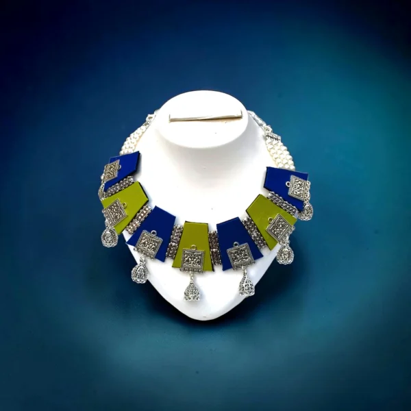 Handmade blue and green fabric necklace set with oxidized silver charms displayed on a white stand against a dark blue background.