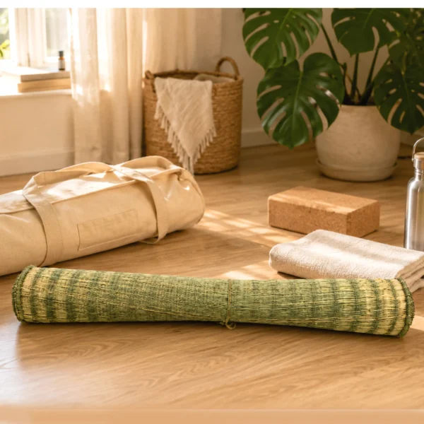 Rolled handmade natural reed grass yoga and meditation mat on a wooden floor with a yoga bag, cork block, towel, and water bottle, highlighting its lightweight, portable, eco-friendly design.
