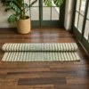 “Handmade natural grass mat with green and cream striped weave, partially rolled on a dark wooden floor beside an indoor plant and glass doors, showing its natural texture and hand-finished edges.”