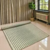 “Handmade natural grass mat with green and cream stripes, fully spread on a polished floor with one end rolled up. The large woven mat has a natural textured surface and hand-finished edges in a bright modern room with curtains, an indoor plant and a window.”